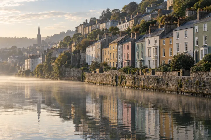 Painted terraced houses stepping up a hill above a misty river at dawn, in a city like Cork.